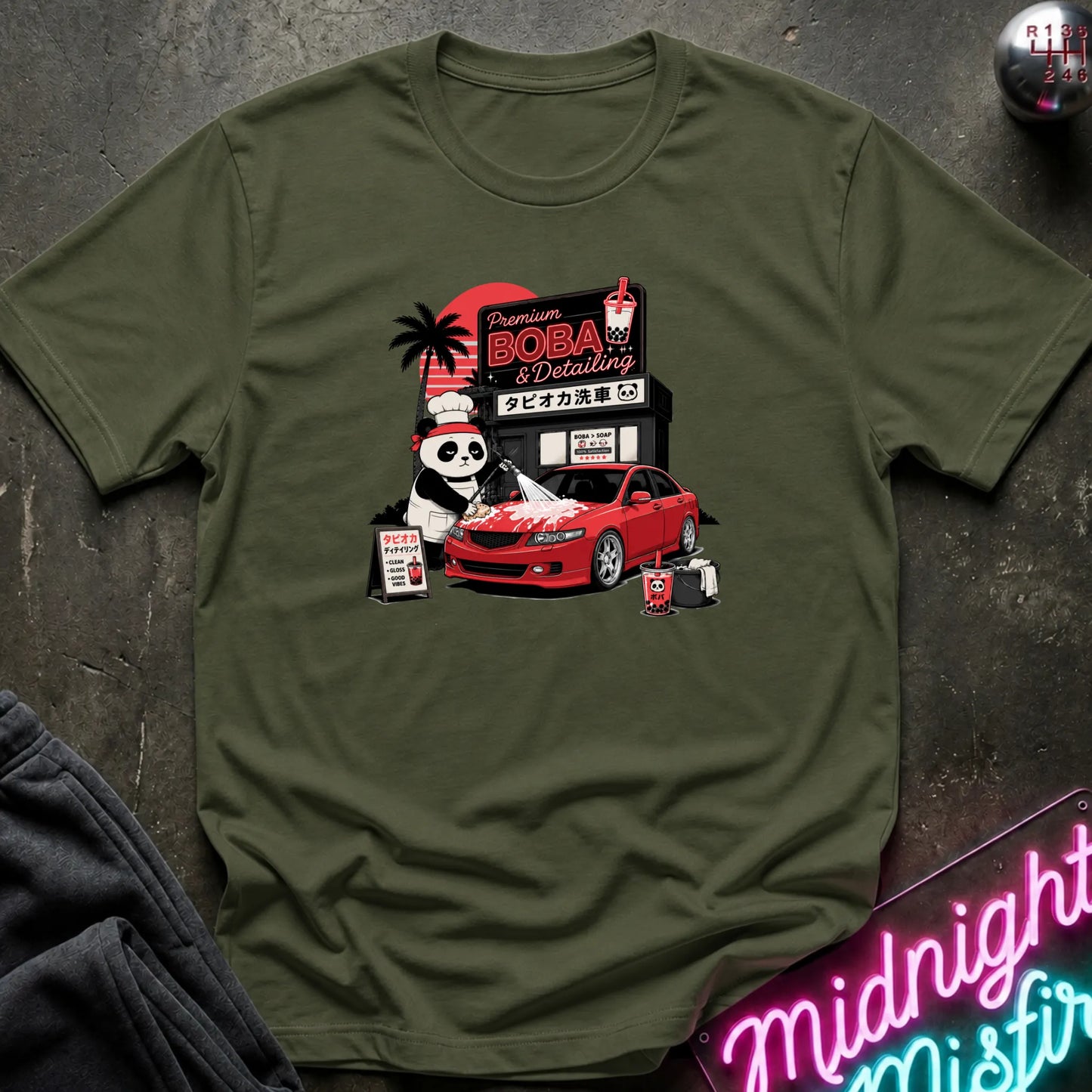 An olive green short-sleeve t-shirt with a detailed graphic print of a panda working at a car detailing shop next to a red sports car, laid flat on a dark textured surface.
