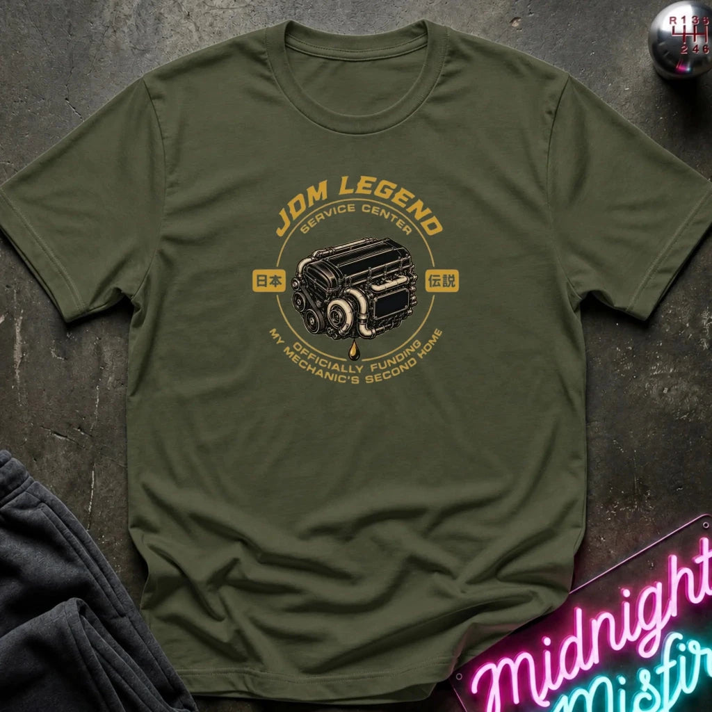 olive graphic t-shirt laid flat on a dark textured surface. The shirt features a vintage-style yellow and black engine graphic with the text 'JDM LEGEND' and 'MY SPECIALTY FUNDS MECHANIC'S SECOND HAND'.