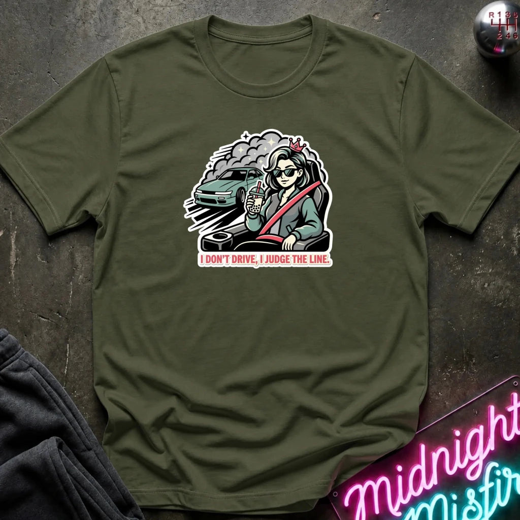 An olive green short-sleeve t-shirt laid flat on a dark textured surface, featuring a graphic of a woman in a car with the text 'I DON'T DRIVE, I JUDGE THE LINE.' A neon sign and folded dark fabric are partially visible around the shirt.