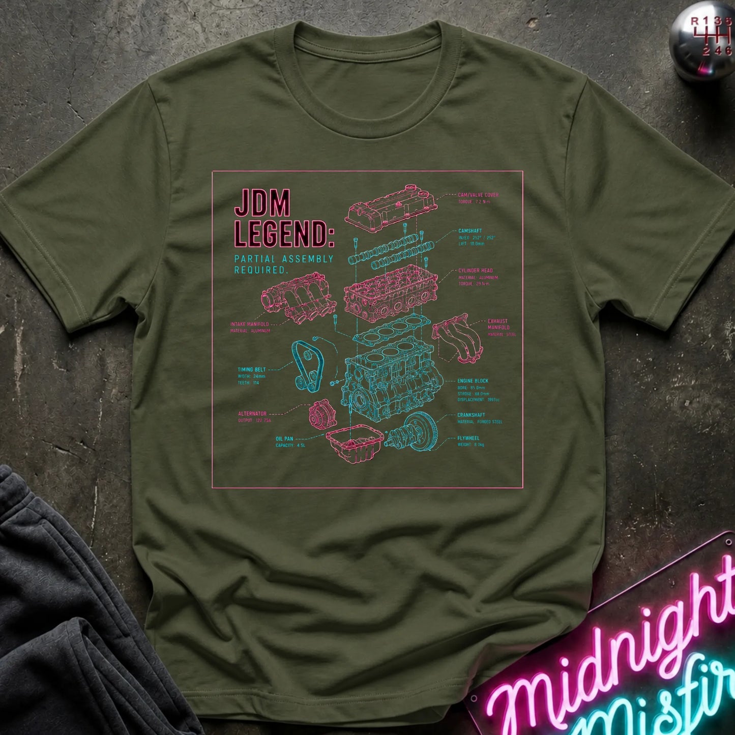 An olive green short-sleeve t-shirt laid flat on a dark textured surface, featuring a 'JDM LEGEND' graphic with pink and teal exploded-view illustrations of a car engine. 