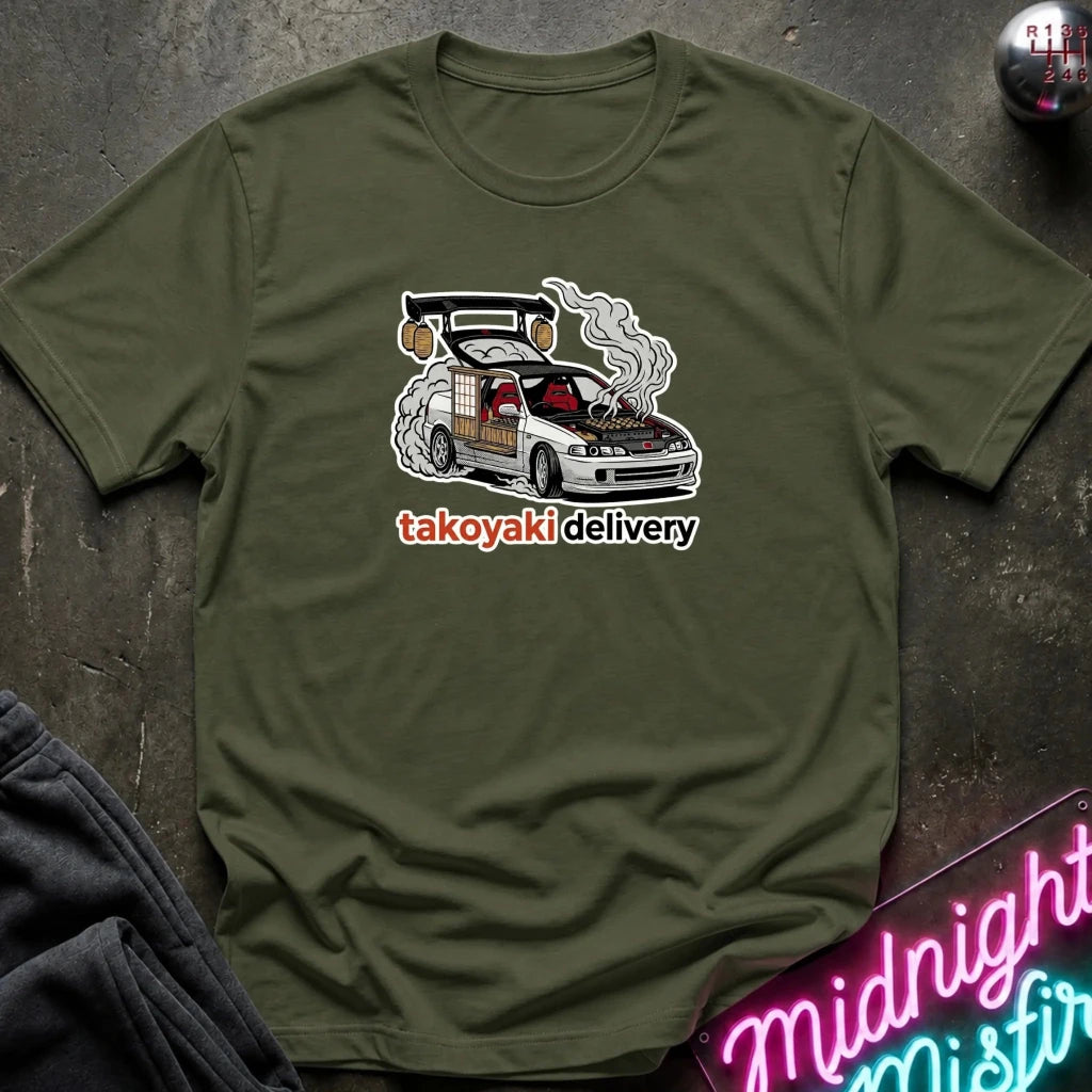 An olive green crewneck t-shirt laid flat on a dark textured surface, featuring a graphic of a white car with its hood and trunk open, smoking, above the text 'takoyaki delivery' in white with a red outline.