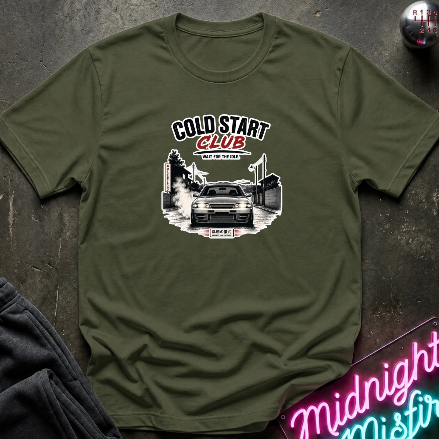 An olive green short-sleeve t-shirt with a 'Cold Start Club' car graphic, laid flat on a dark textured surface with a neon sign and dark pants partially visible.