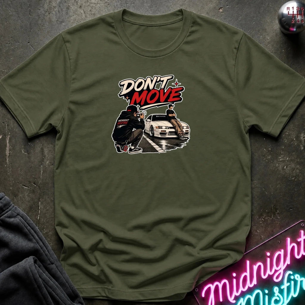 An olive green short-sleeve t-shirt with a graphic print on the chest that reads 'DON'T MOVE' above an illustration of a person and a car. The shirt is laid flat on a dark textured surface with a neon sign and dark pants partially visible in the corners.