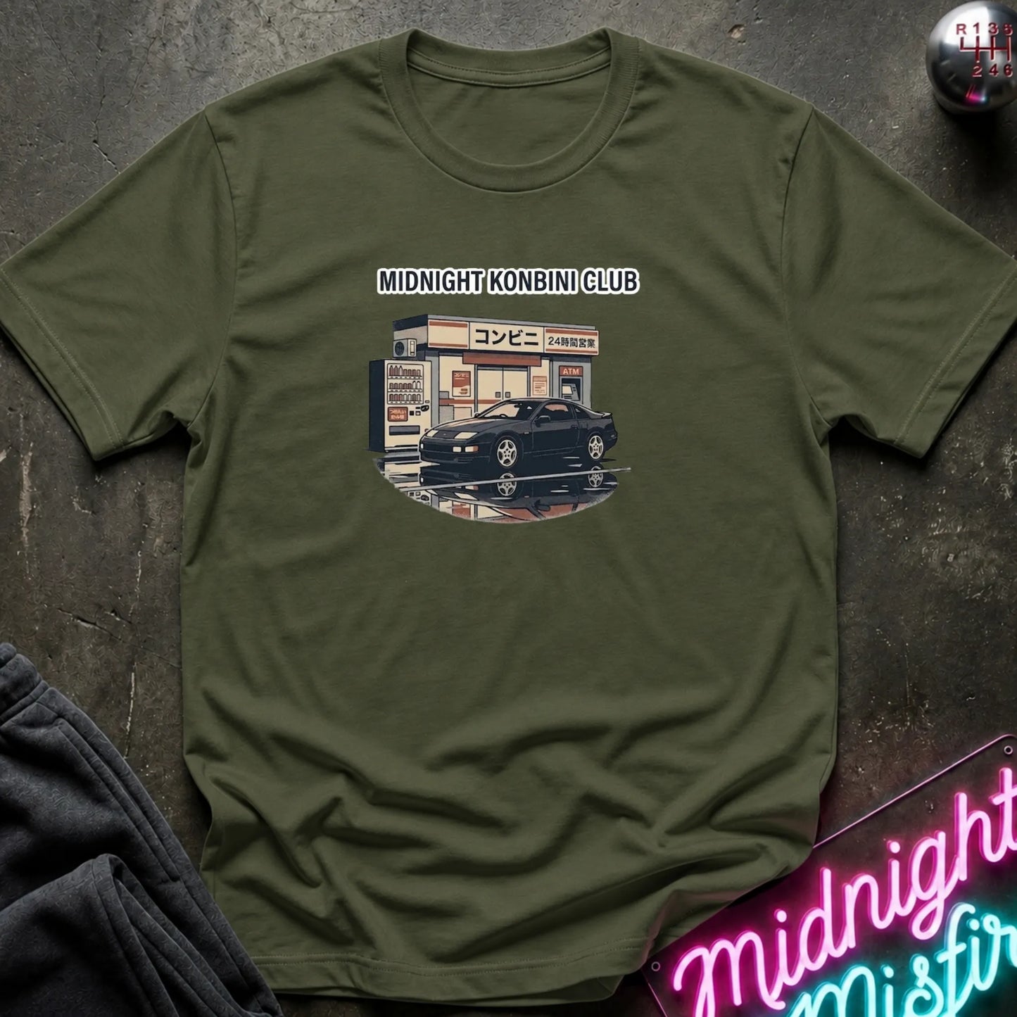 An olive green short-sleeve t-shirt with a graphic print of a Japanese convenience store and a black sports car under the text 'MIDNIGHT KONBINI CLUB'. The shirt is laid flat on a dark textured surface with a neon sign and dark pants partially visible.