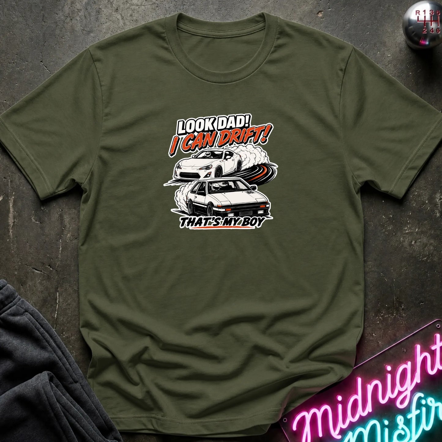 An olive green  short-sleeved t-shirt  that has a cars drifting which are older and newer generations of toyotas.