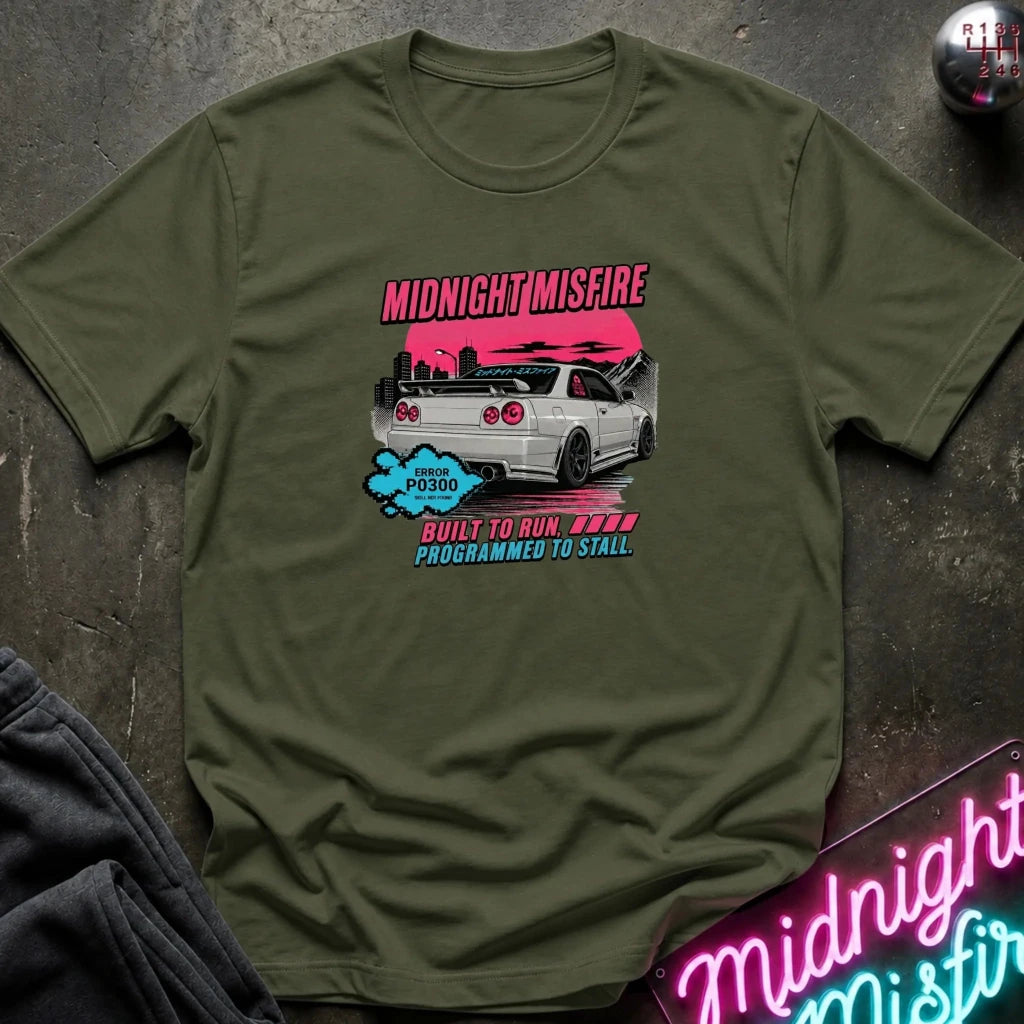 An olive green graphic t-shirt laid flat on a dark textured surface. The graphic features a white Nissan S350 R33 with a pink sunset sky and the text 'MIDNIGHTMISFIRE' and 'BUILT TO RUN PROGRAMMED TO STALL'.