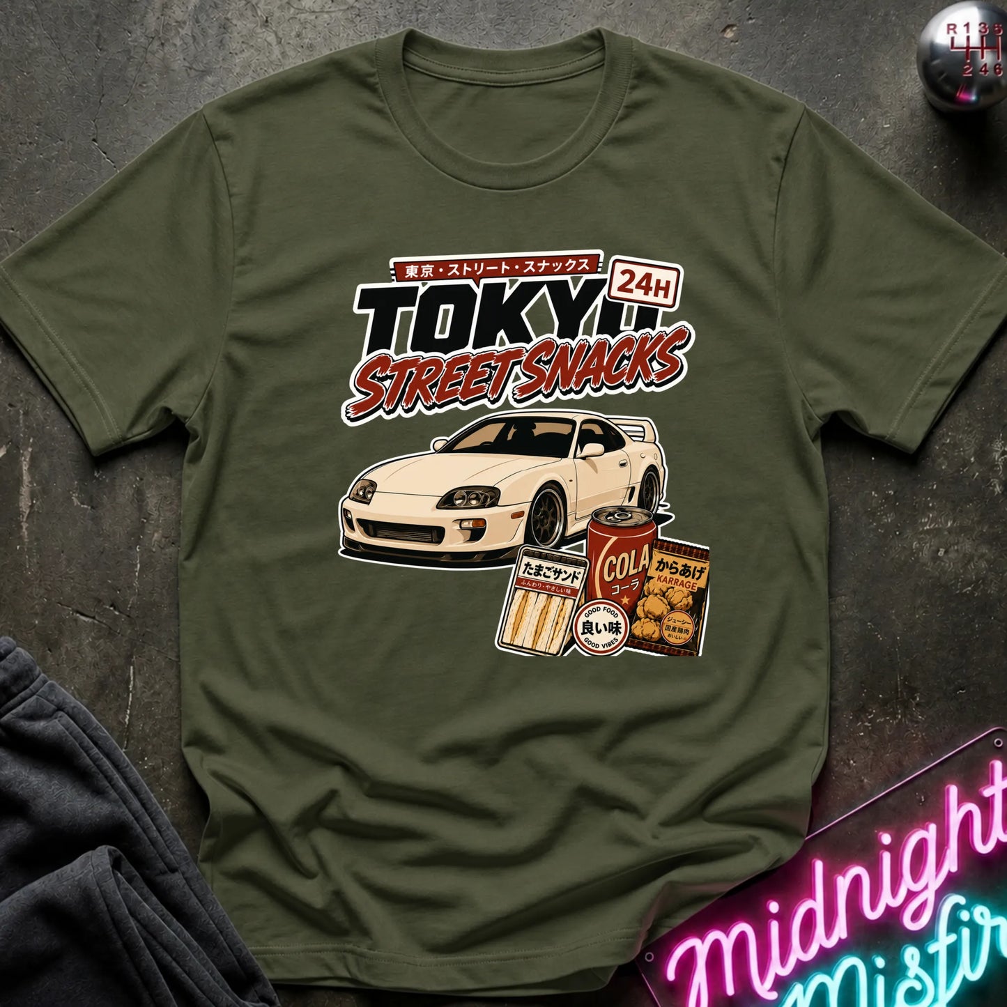 An olive green graphic t-shirt laid flat on a dark textured surface. The graphic features a white sports car and retro Japanese-style snack cans with the text 'TOKYO STREET SNACKS 24H'.