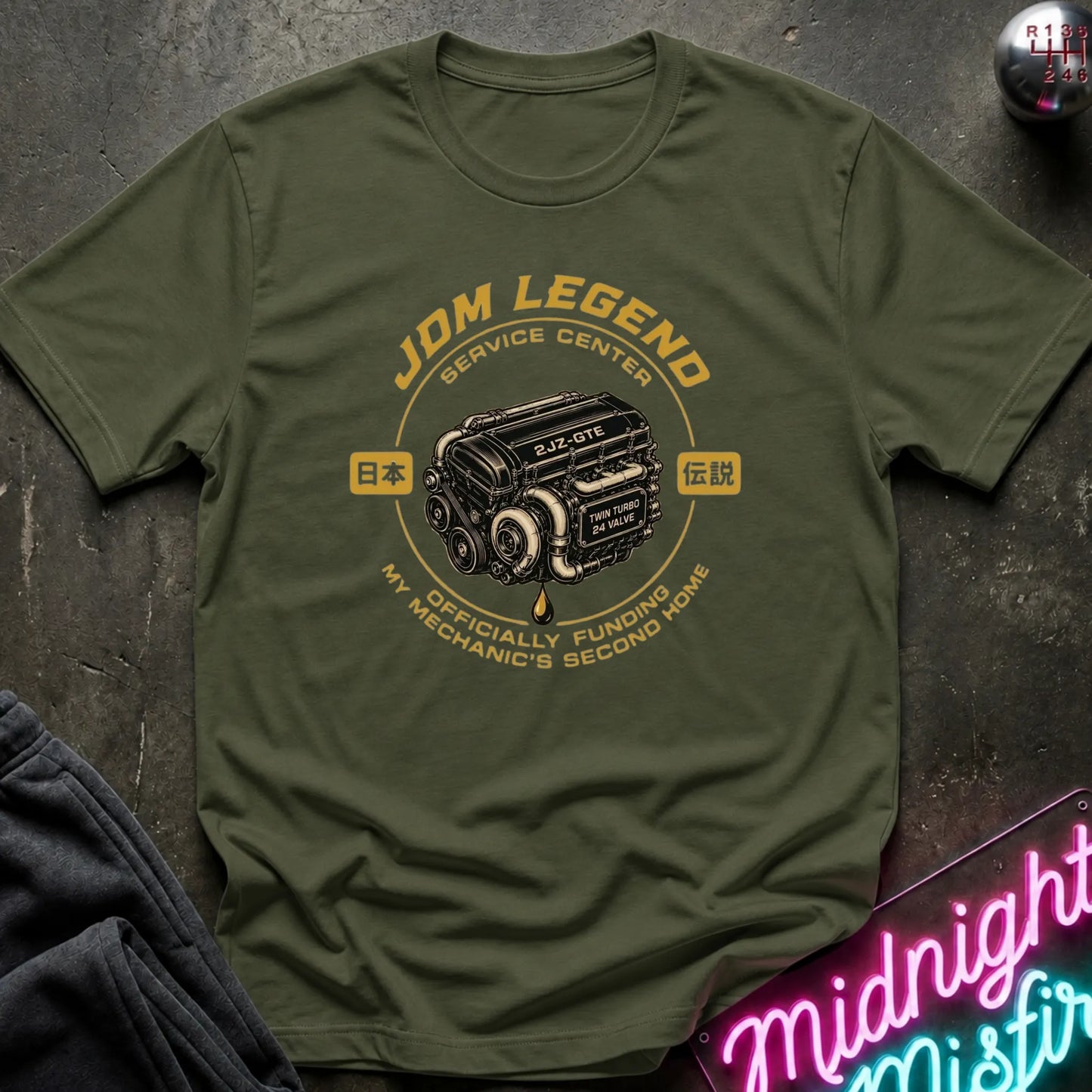 An olive green short-sleeve t-shirt laid flat on a dark textured surface. The shirt features a vintage-style graphic of a Japanese car engine with the text 'JDM LEGEND SERVICE CENTER' and 'MY MECHANIC'S SECOND HOME' in yellow and white.