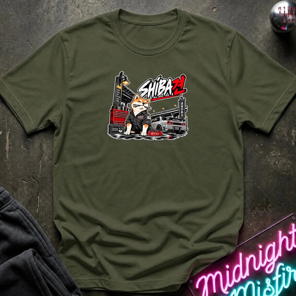 An olive green crewneck t-shirt featuring a graphic print of a Shiba Inu character with a tool chest and the text 'SHIBA'. The shirt is displayed flat on a dark textured surface with a neon sign and dark sweatpants partially visible.
