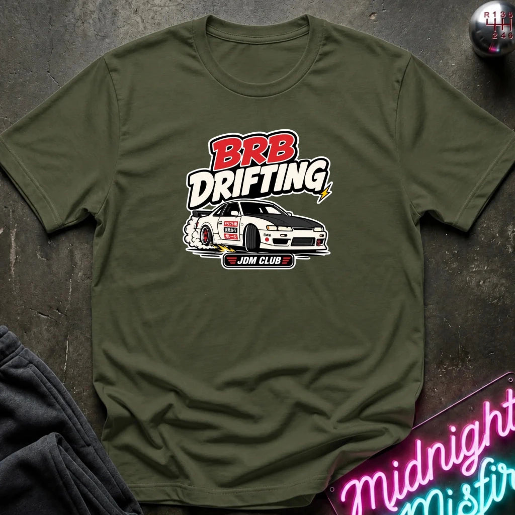 An olive green short-sleeve t-shirt with a 'BRB Drifting ' graphic featuring a white drift car, laid flat on a dark textured surface with a neon sign and sweatpants partially visible.