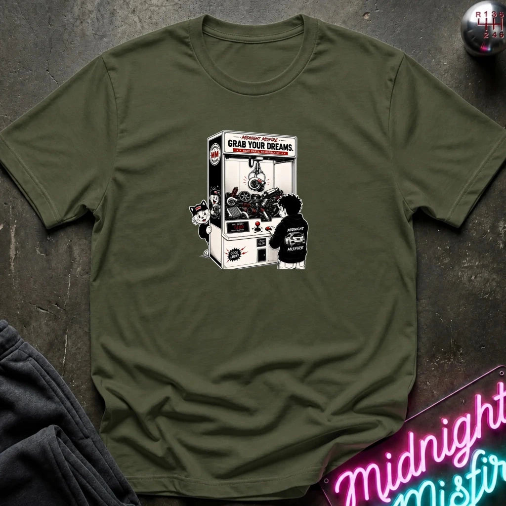 An olive green short-sleeve t-shirt laid flat on a dark textured surface, featuring a black and white graphic print of a claw machine with the text 'GRAB YOUR DREAMS'. 