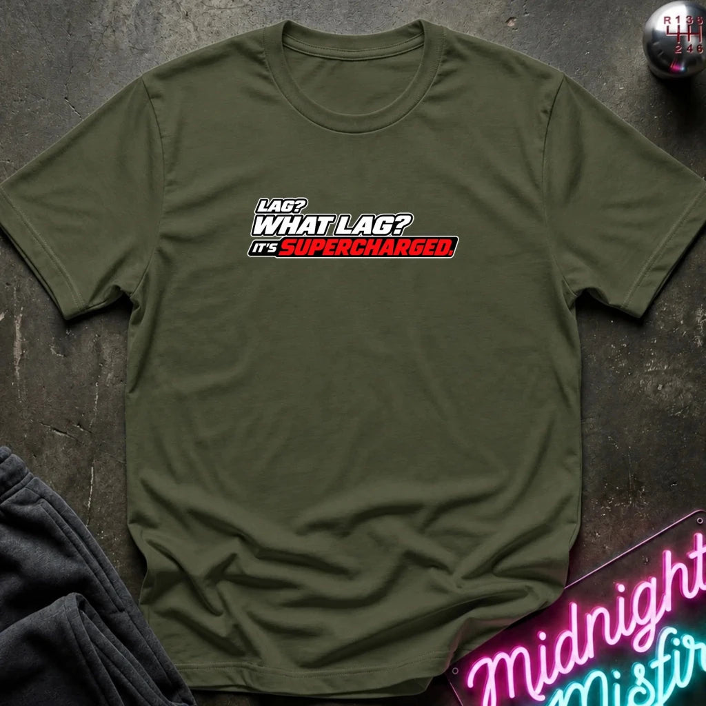An olive green short-sleeve t-shirt with a graphic text print reading 'LAG? WHAT LAG? IT'S SUPERCHARGED' in white, black, and red. The shirt is displayed flat on a dark textured surface with a neon sign and dark pants partially visible as props.
