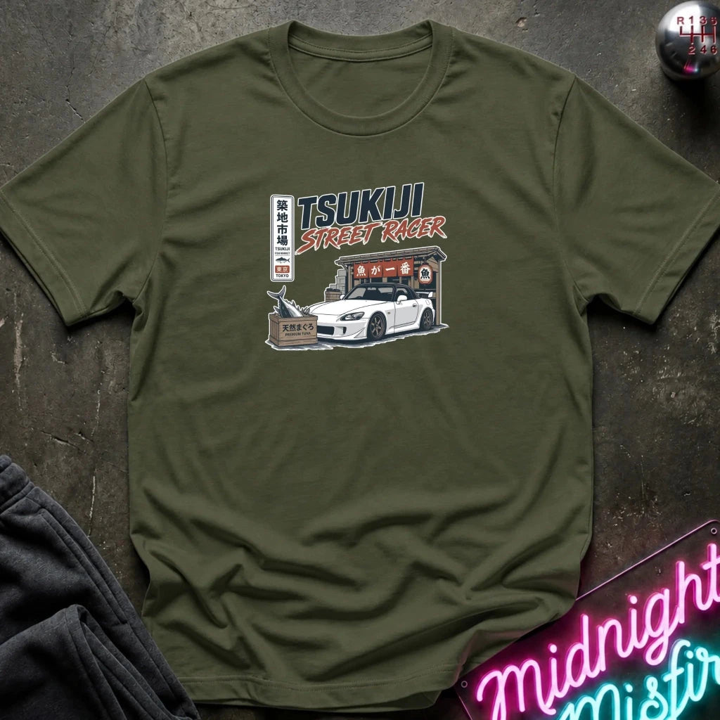 An olive green crewneck t-shirt featuring a graphic print of a white sports car and Japanese text reading 'TSUKIJI STREET RACER'. The shirt is styled flat on a dark textured surface with a neon sign and dark pants partially visible.