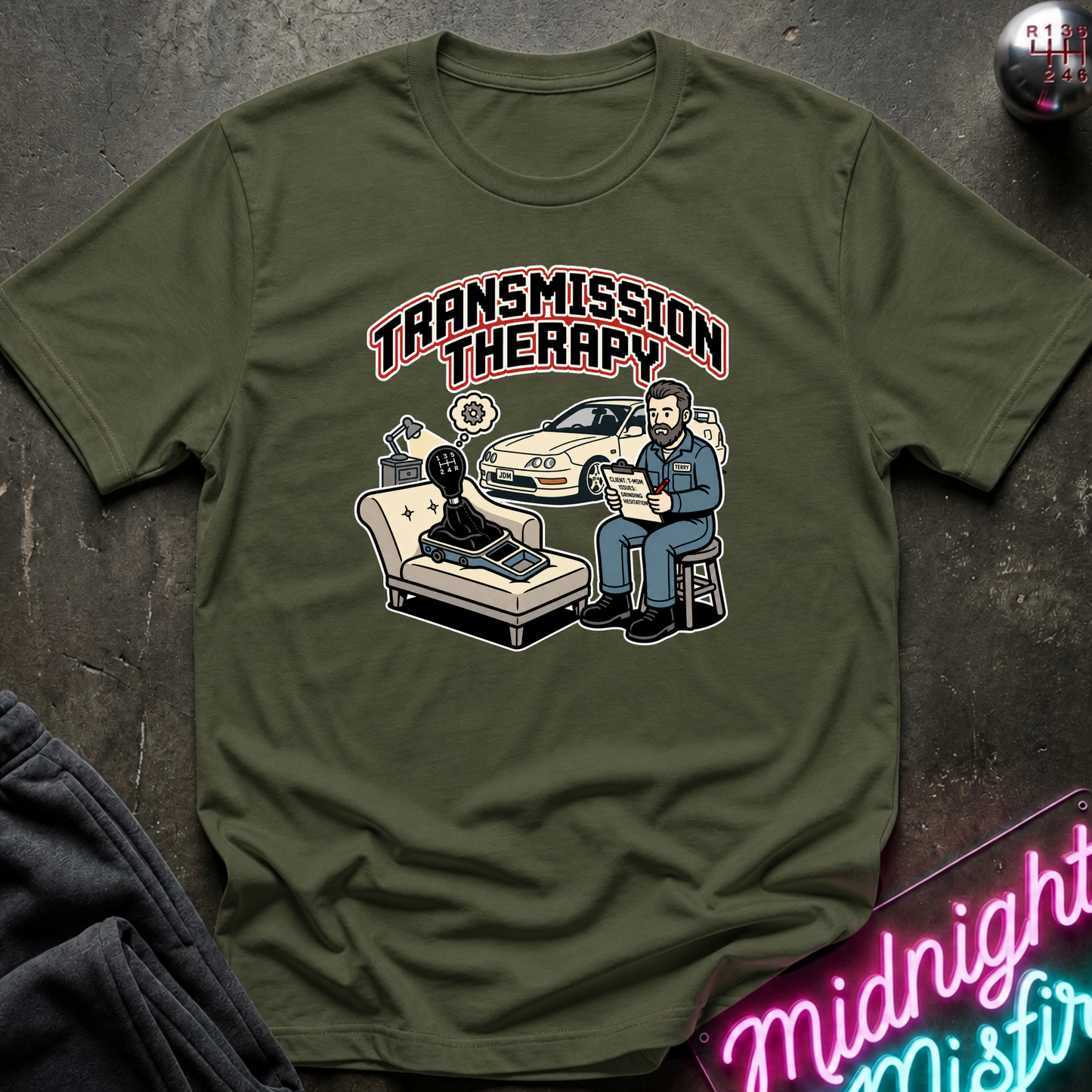 An olive green graphic t-shirt laid flat on a dark textured surface. The graphic features the words 'TRANSMISSION THERAPY' in arched pink and white lettering above a cartoon illustration of a therapist talking to a client in a chair, with a car in the background.