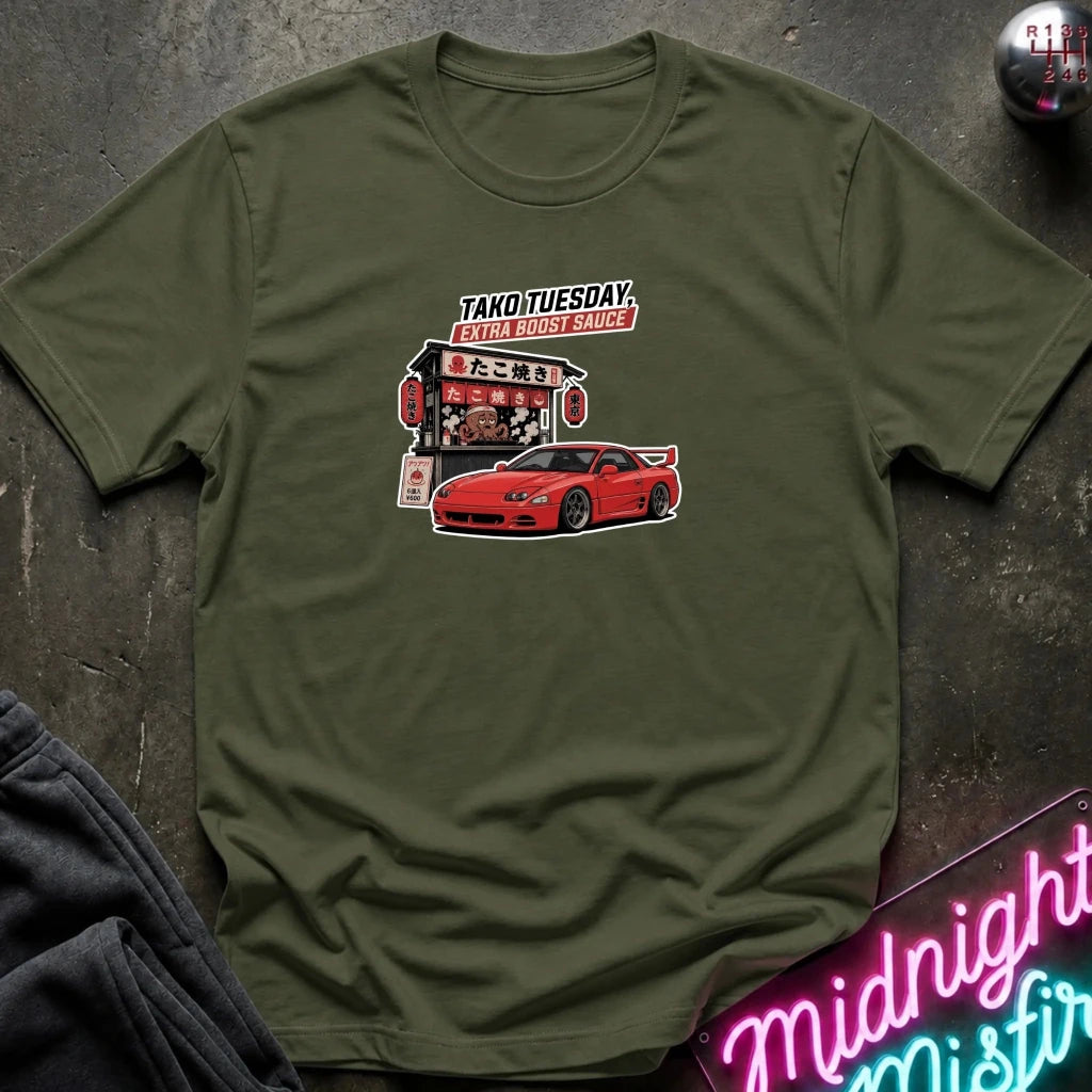 An olive green short-sleeve t-shirt with a graphic print of a red sports car and a Japanese food stall under the text 'TAKOTUESDAY', laid flat on a dark textured surface with a neon sign and dark pants partially visible.