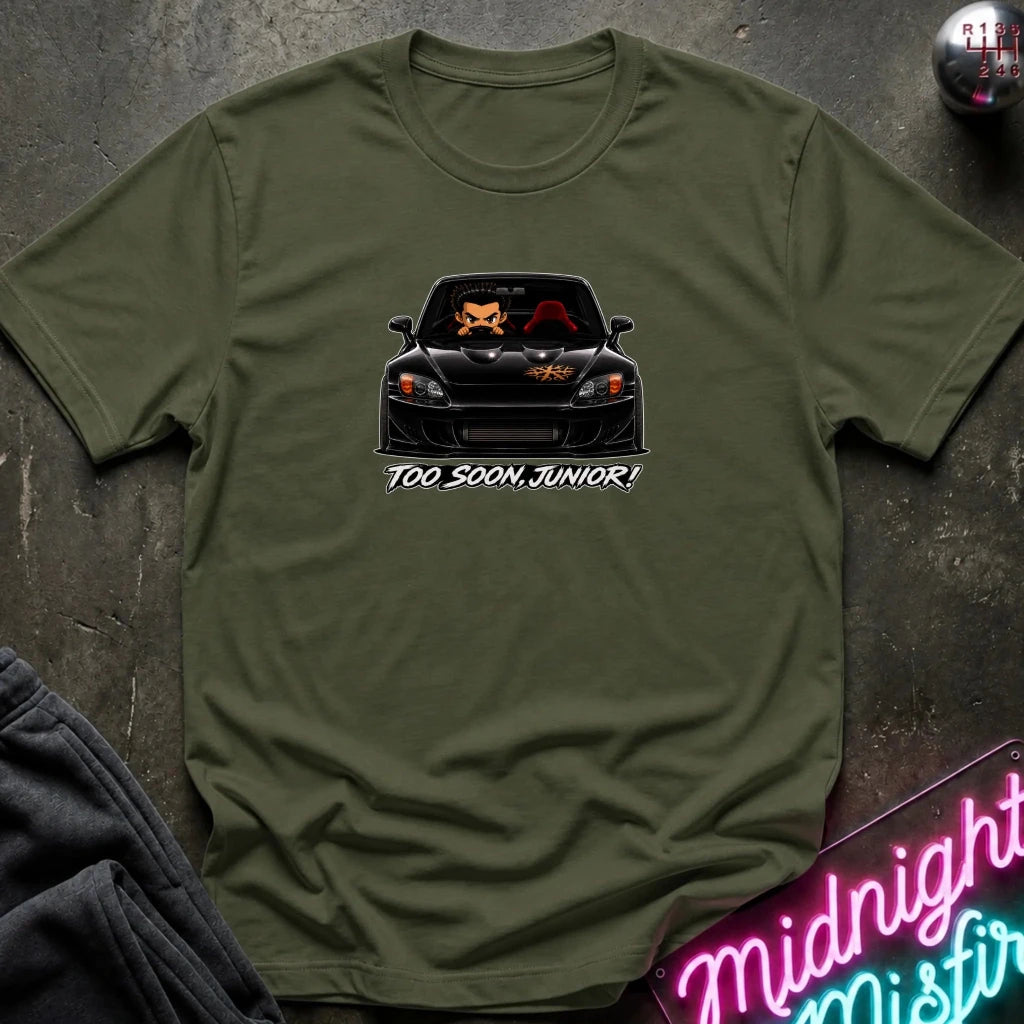 An olive green short-sleeve t-shirt laid flat on a dark textured surface, featuring a graphic of a black sports car with orange foxes in the windshield and the text 'TOO SOON, JUNIOR!' below. A neon sign and folded dark fabric are partially visible in the corners.