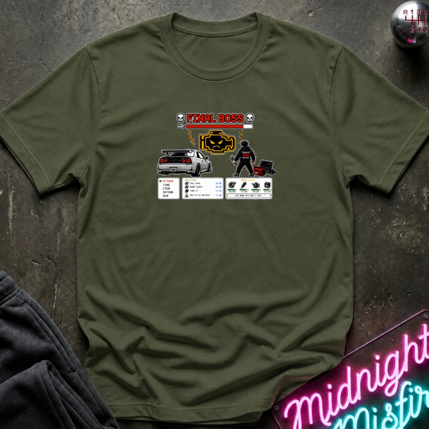 A olive green short-sleeve crewneck t-shirt laid flat on a dark textured surface, featuring a central graphic print of a rpg character and a car. 