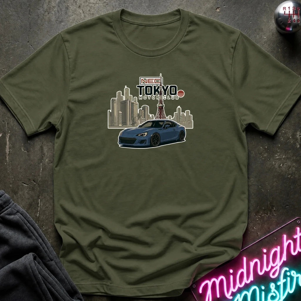 An olive green crewneck t-shirt laid flat on a dark textured surface, featuring a graphic print of a blue sports car in front of a Tokyo city skyline
