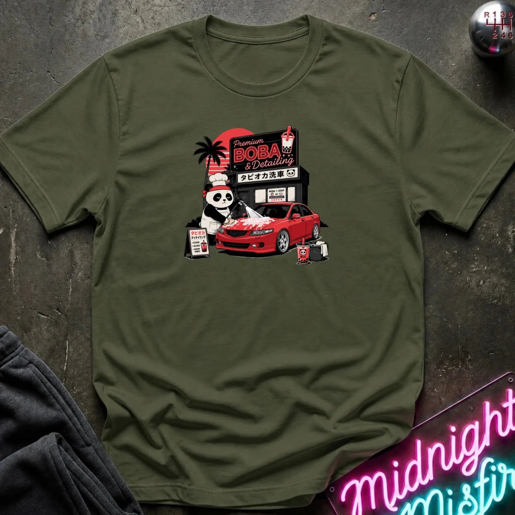 olive green short-sleeve graphic t-shirt laid flat on a dark textured surface. The graphic features a cartoon panda wearing a hat next to a red sports car, with a sign reading 'Midnight Boba'.