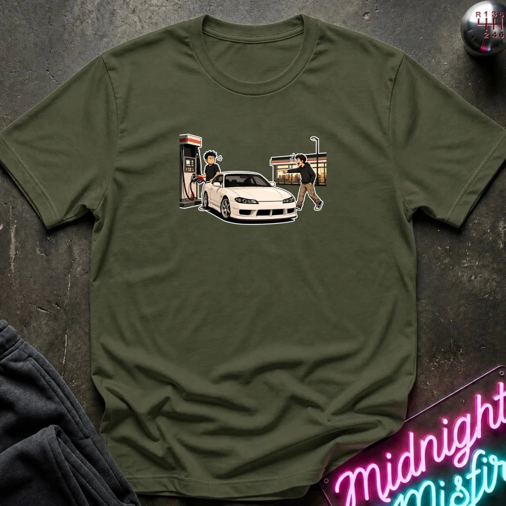 An olive green short-sleeve t-shirt with a graphic print of a white sports car at a gas station, laid flat on a dark textured surface with a neon sign and dark pants partially visible.