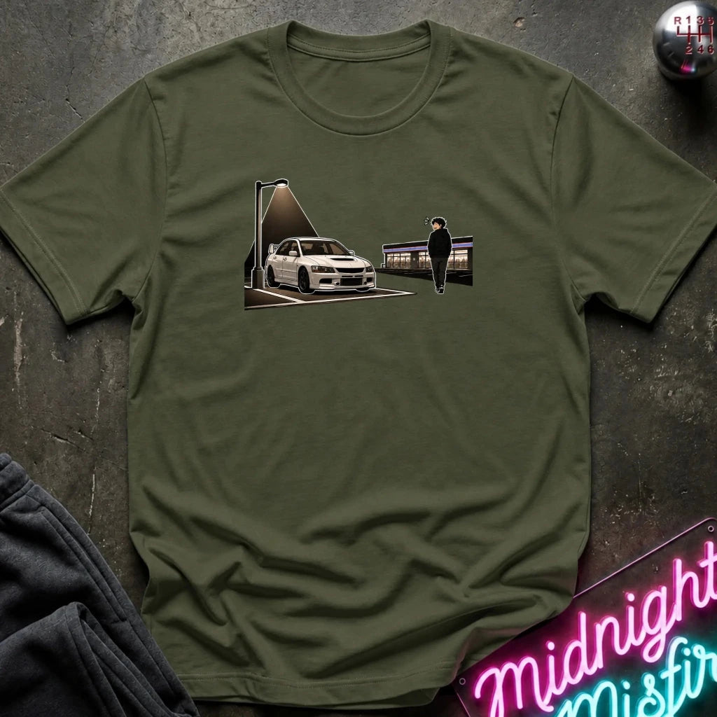 An olive green crewneck t-shirt laid flat on a dark textured surface, featuring a graphic print of a white sports car and a person under a streetlamp. Styled flat-lay props include a neon sign in the corner and a pair of dark jeans.