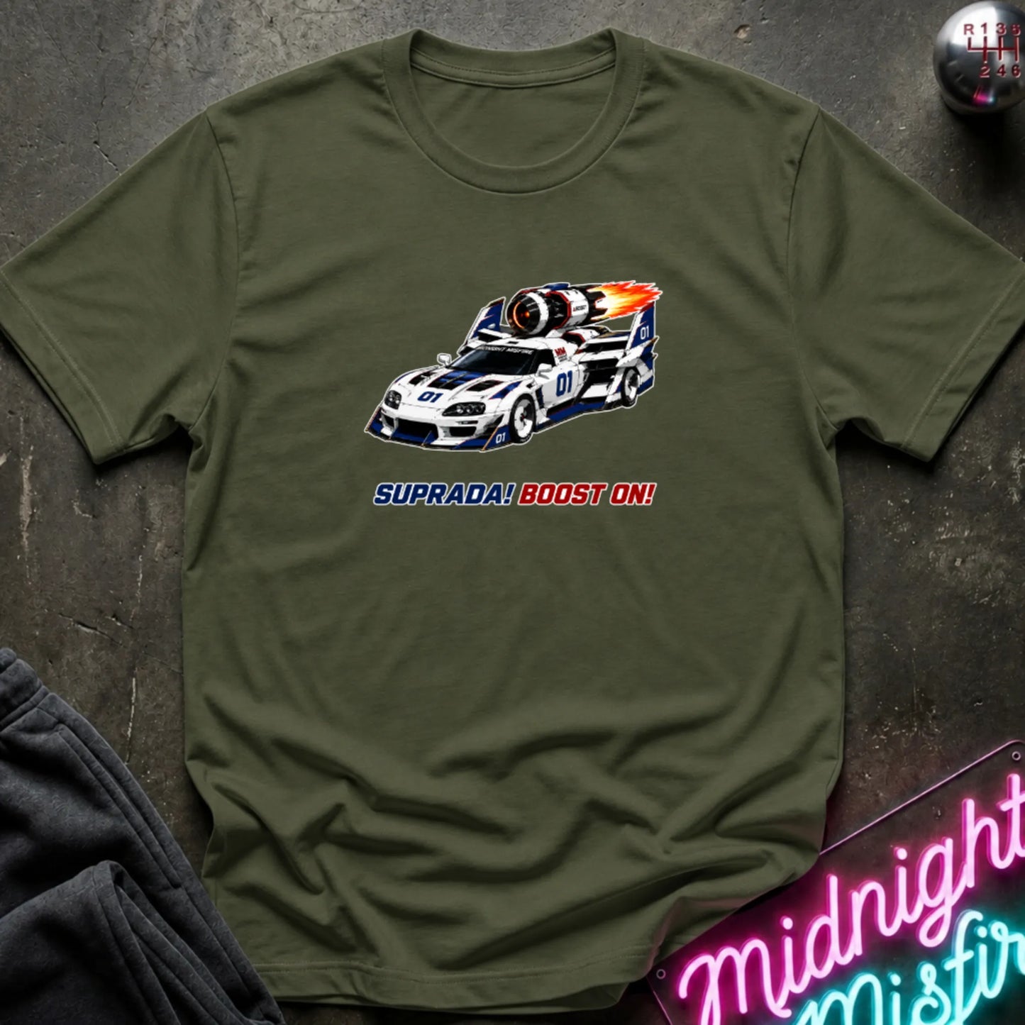  A olive green short-sleeve t-shirt with a retro-style graphic of a race machine with big boost, displayed flat on a dark textured surface with a neon sign and sweatpants partially visible.