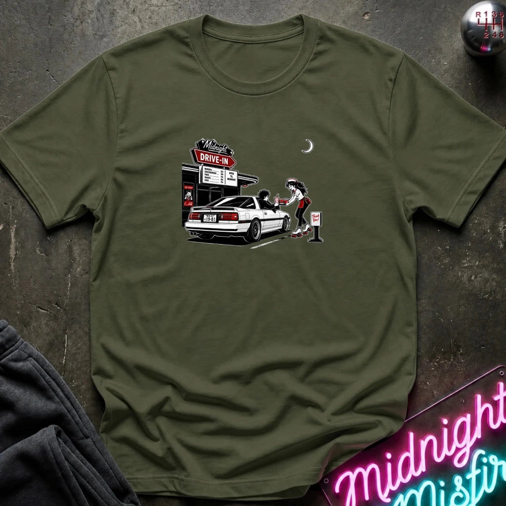 A olive green short-sleeve t-shirt laid flat on a dark textured surface, featuring a graphic print of a woman pumping gas at a drive-in station next to a white car. 