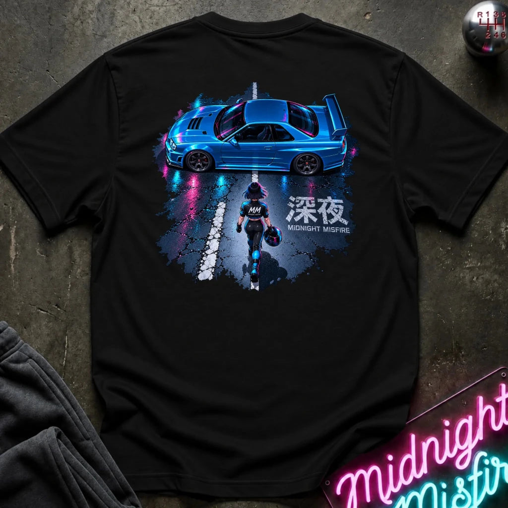 A black short-sleeve t-shirt laid flat on a dark textured surface, featuring a large graphic print of a blue sports car and a motorcycle rider with Japanese text. 