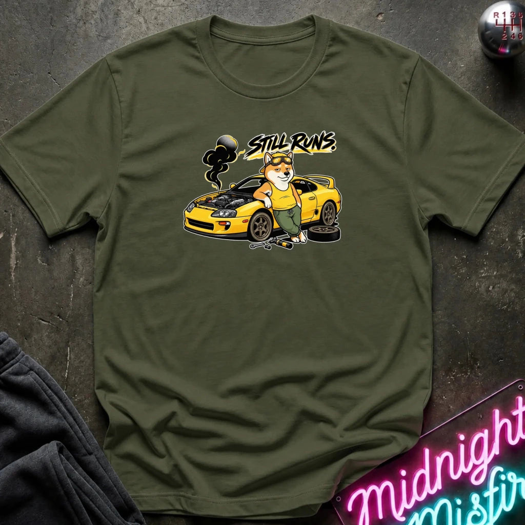 olive short-sleeve graphic t-shirt laid flat on a dark textured surface. The graphic features a cartoon dog in a mechanic hat standing next to a yellow sports car with the text 'Still runs' above it.
