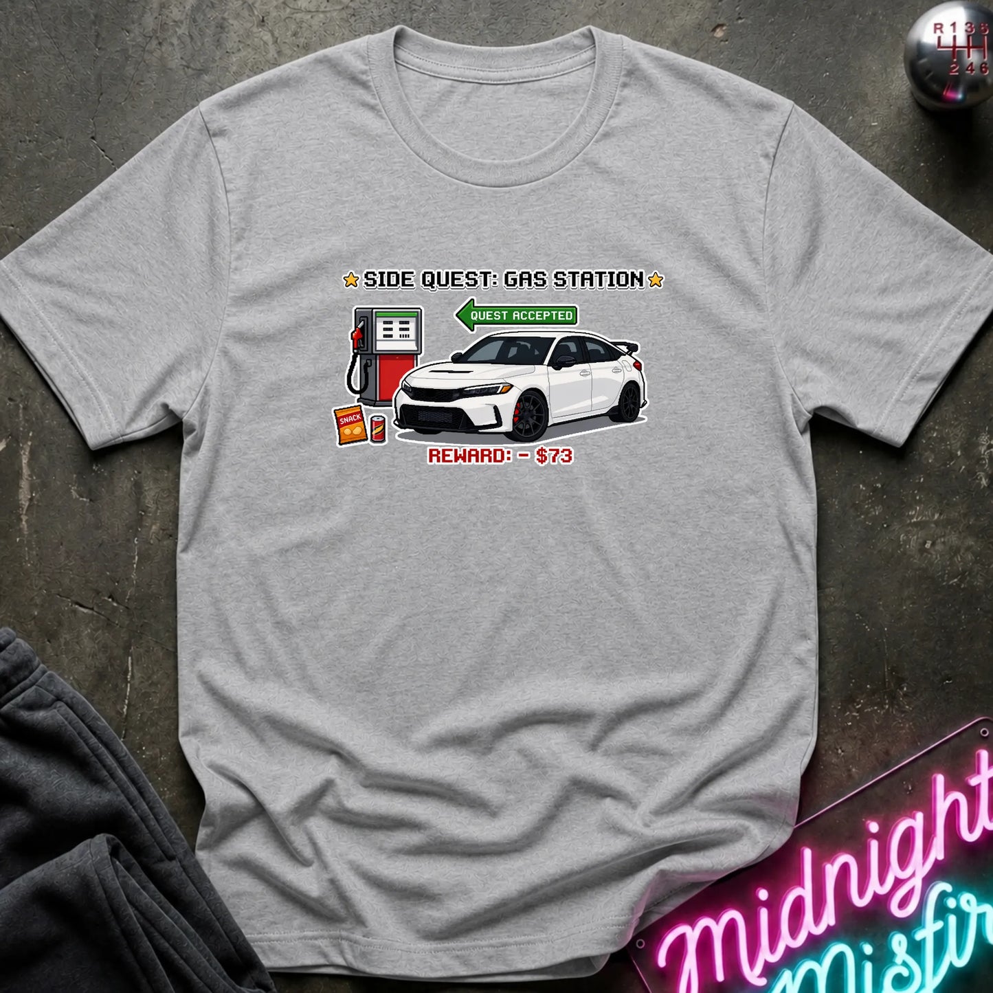 A light gray heather crewneck t-shirt featuring a graphic print of a white sports car at a gas station with the text 'SIDE QUEST: GAS STATION' and 'REWARD: $73'. The shirt is laid flat on a dark textured surface with a neon sign and dark pants partially visible in the corners.