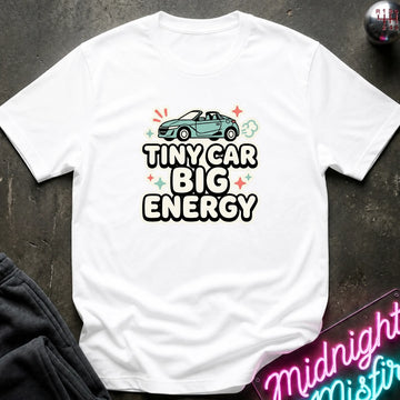 A white short-sleeve graphic T-shirt laid flat on a dark textured surface. The shirt features a light blue convertible car illustration above the text 'TINY CAR BIG ENERGY' in bold black letters with white outlines, accented by small colorful star and cloud shapes.