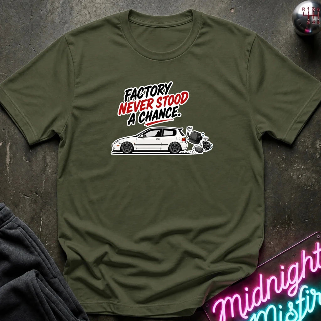 An olive green short-sleeve t-shirt with a graphic print of a white car and the text 'FACTORY NEVER STOOD A CHANCE.' in black and red, laid flat on a dark textured surface.