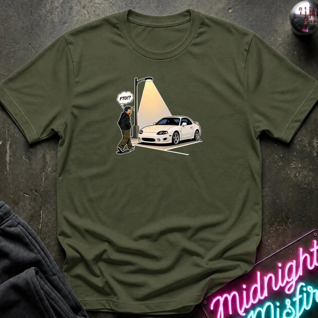 An olive green crewneck t-shirt laid flat on a dark textured surface, featuring a graphic print of a white sports car parked in a spot with a streetlamp shining on it and a cartoon figure with a speech bubble 