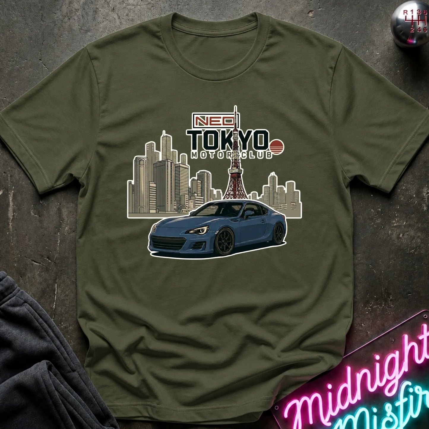 An olive green short-sleeve t-shirt with a graphic print of a blue sports car and the text 'NEO TOKYO MOTOR CLUB' against a city skyline. The shirt is displayed flat on a dark textured surface with a neon sign and dark sweatpants partially visible.