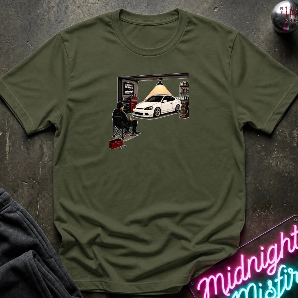 An olive green crewneck t-shirt laid flat on a dark textured surface, featuring a graphic print of a white sports car in a garage with a person sitting in a folding chair. A neon sign and folded dark fabric are partially visible in the corners.