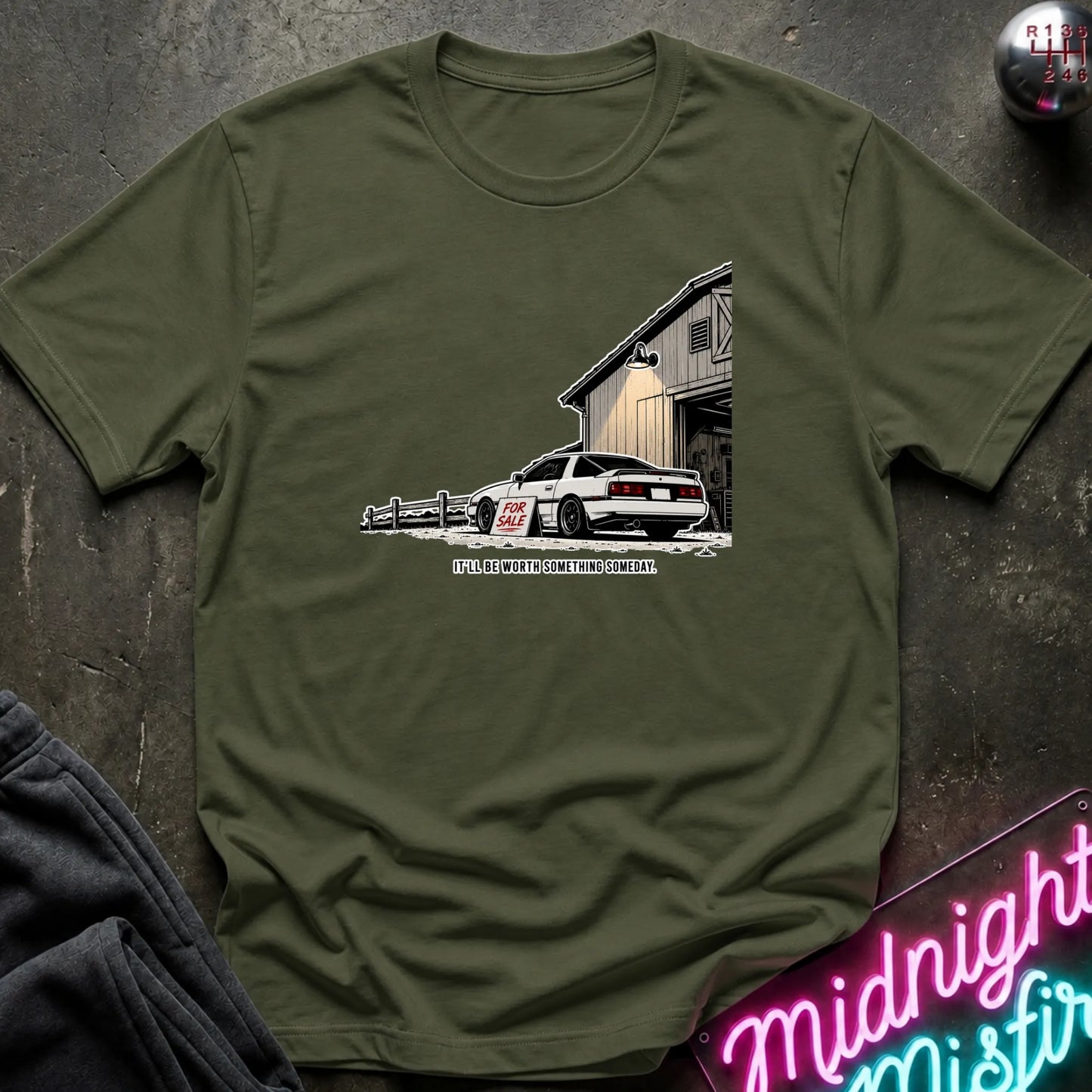 An olive green crewneck t-shirt laid flat on a dark textured surface, featuring a graphic print of a white sports car parked in front of a barn with the text.