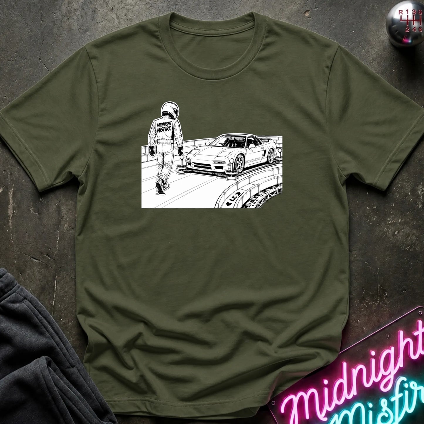 An olive green crewneck t-shirt featuring a black and white graphic print of a race car and a driver, laid flat on a dark textured surface with a neon sign and dark sweatpants nearby.