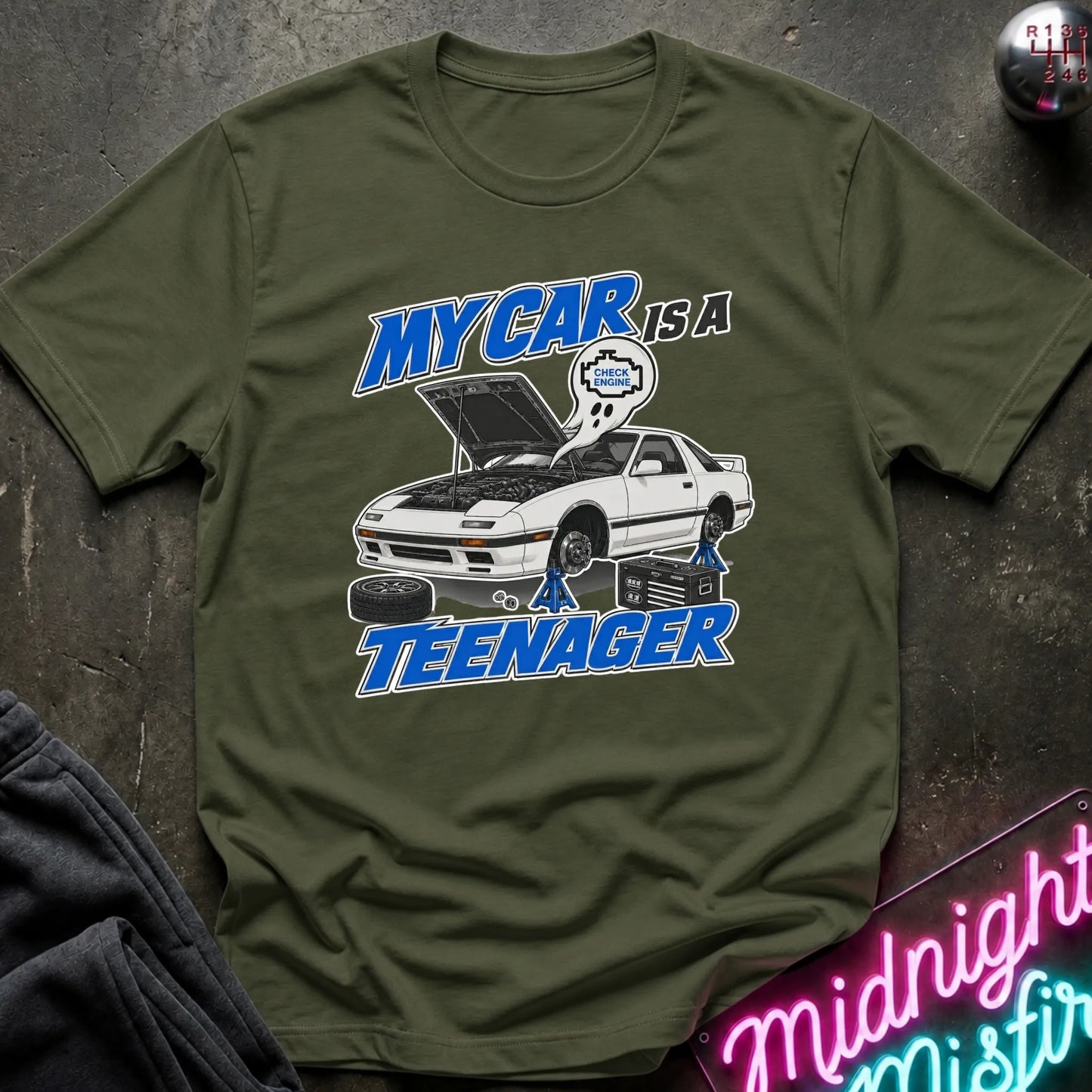 An olive green graphic t-shirt laid flat on a dark textured surface. The graphic features a white sports car with its hood open, blue text reading 'MY CAR IS A TEENAGER',.
