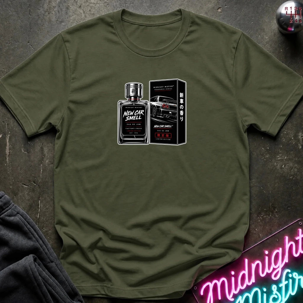 An olive green short-sleeve t-shirt laid flat on a dark textured surface, featuring a graphic print of a perfume bottle and its box labeled 'New Car Smell' with a sports car illustration.