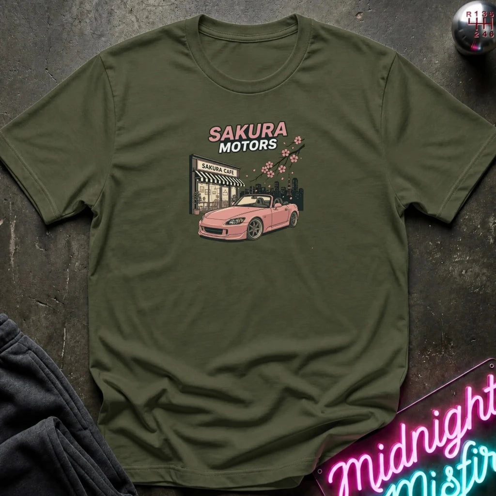 An olive green short-sleeve t-shirt laid flat on a dark textured surface, featuring a graphic print of a pink convertible car in front of a 'Sakura Motors' shop with cherry blossoms. A neon sign and dark clothing are partially visible in the corners as styling props.
