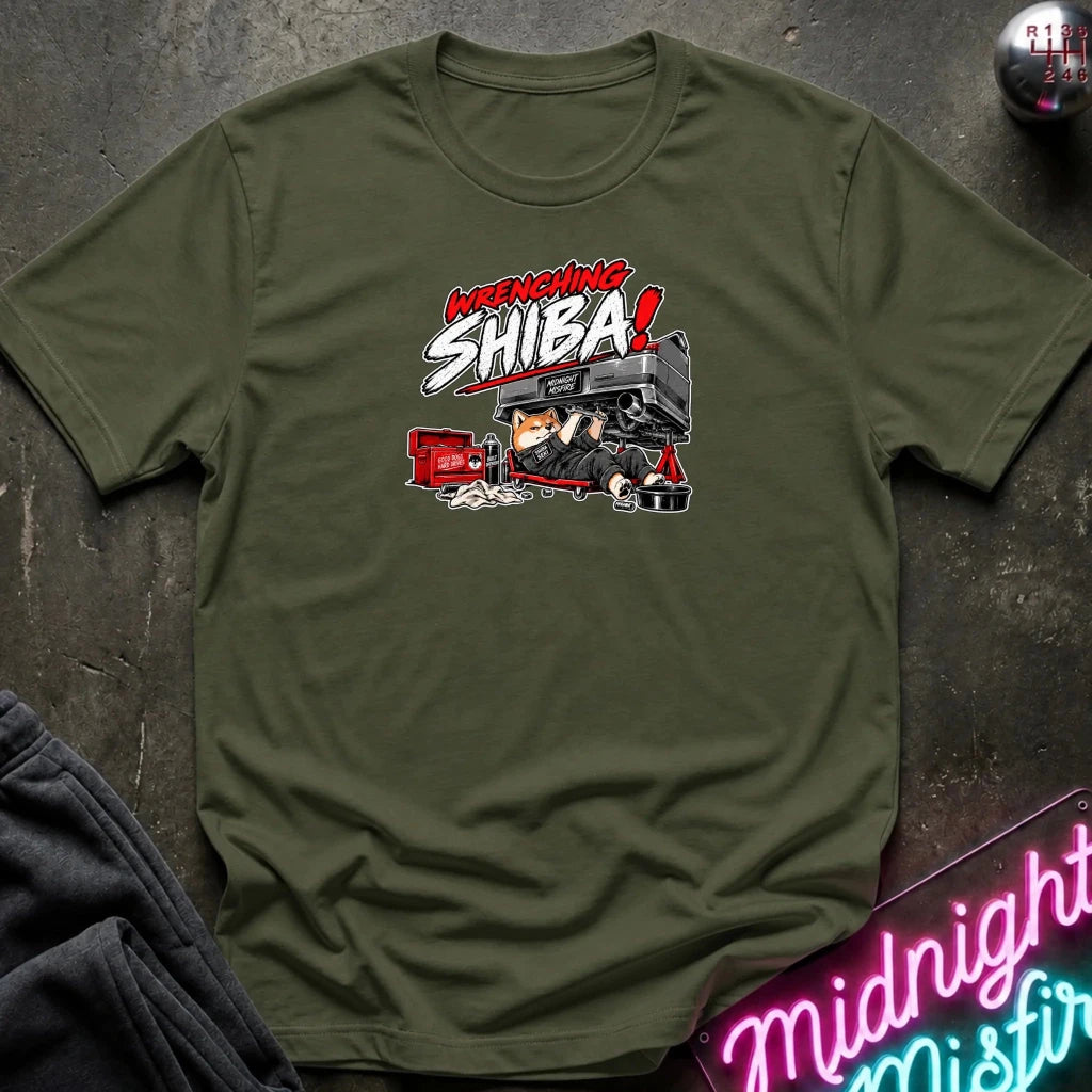 An olive green short-sleeve graphic t-shirt laid flat on a dark textured surface. The graphic features the text 'WRENCHING SHIBA!' in red and white comic-style lettering above an illustration of a character in a black and red vehicle.
