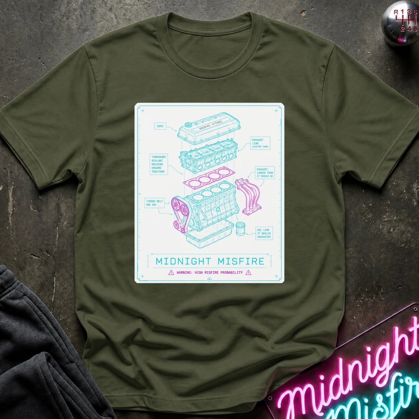 An olive green crewneck t-shirt laid flat on a dark textured surface, featuring a central graphic print of a pink and blue engine diagram labeled 'MIDNIGHT MISFIRE'.