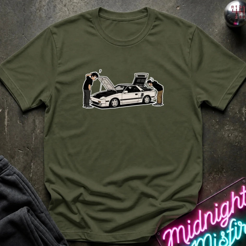 An olive green short-sleeve t-shirt laid flat on a dark textured surface, featuring a graphic print of a white sports car with two people inspecting it. 