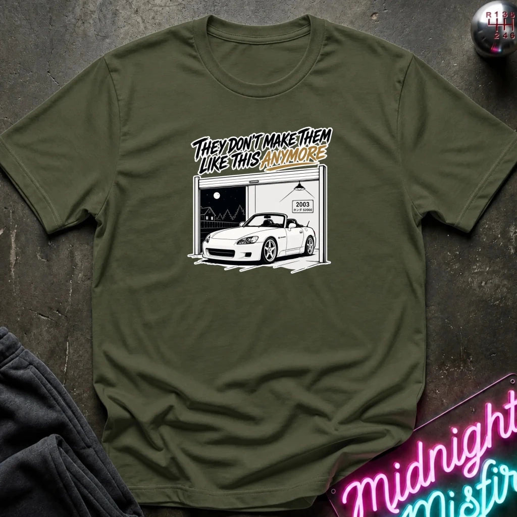 An olive green crewneck t-shirt laid flat on a dark textured surface. The shirt features a graphic print of a white sports car inside a garage with the text 'THEY DON'T MAKE THEM LIKE THIS ANYMORE' in stylized lettering.