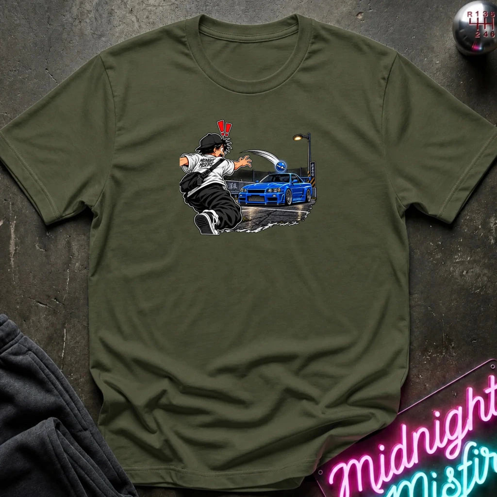An olive green short-sleeve crewneck t-shirt laid flat on a dark textured surface. The center of the shirt features a graphic print of an anime character in a black and white outfit with a hat, next to a blue sports car under a streetlamp.