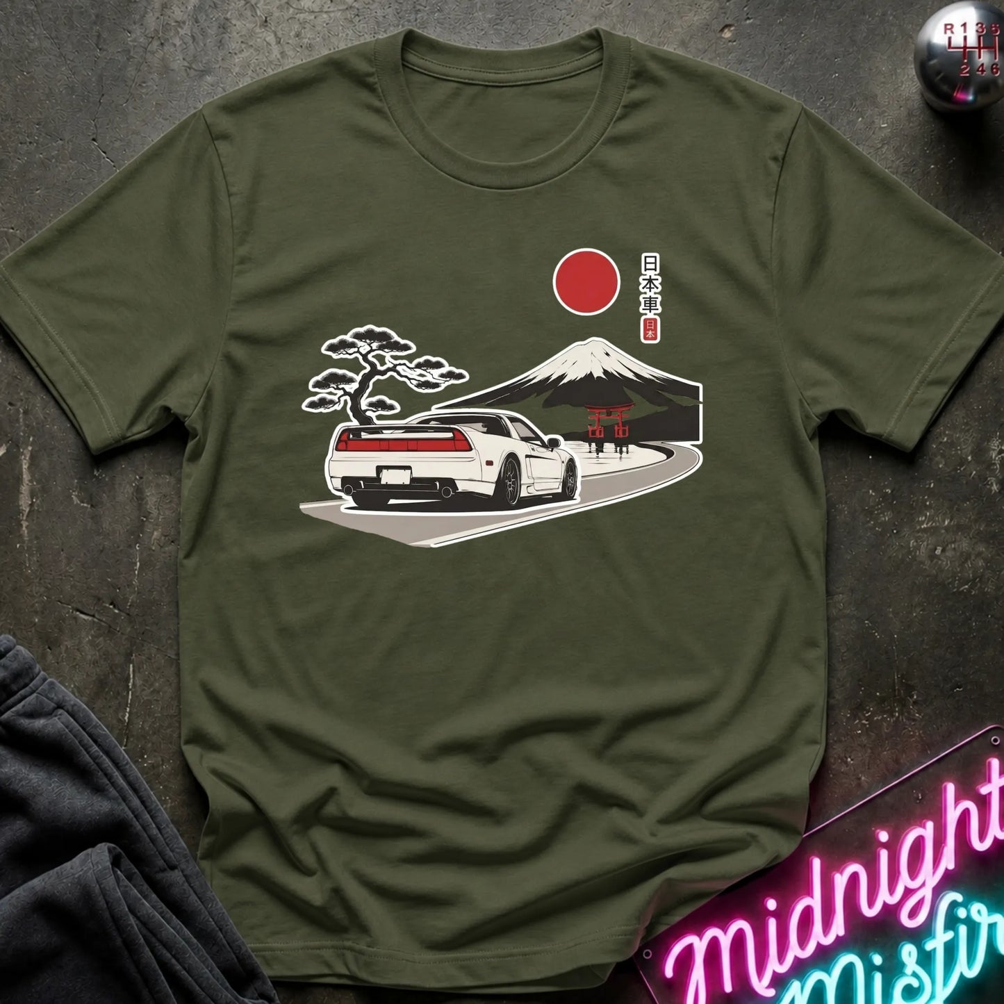 An olive green short-sleeve t-shirt with a graphic print of a white sports car, Mount Fuji, a bonsai tree, a red sun, and Japanese text, displayed flat on a dark textured surface with a neon sign and dark pants partially visible.