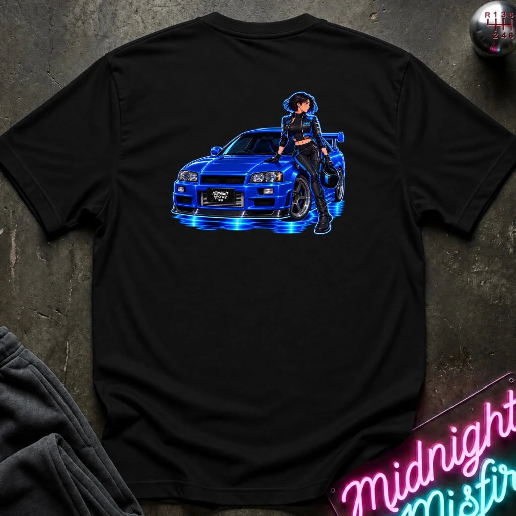 A black short-sleeve t-shirt laid flat on a dark textured surface, featuring a graphic print of a blue sports car with glowing blue lights and an anime-style woman standing beside it. A neon sign and folded dark fabric are partially visible around the shirt.