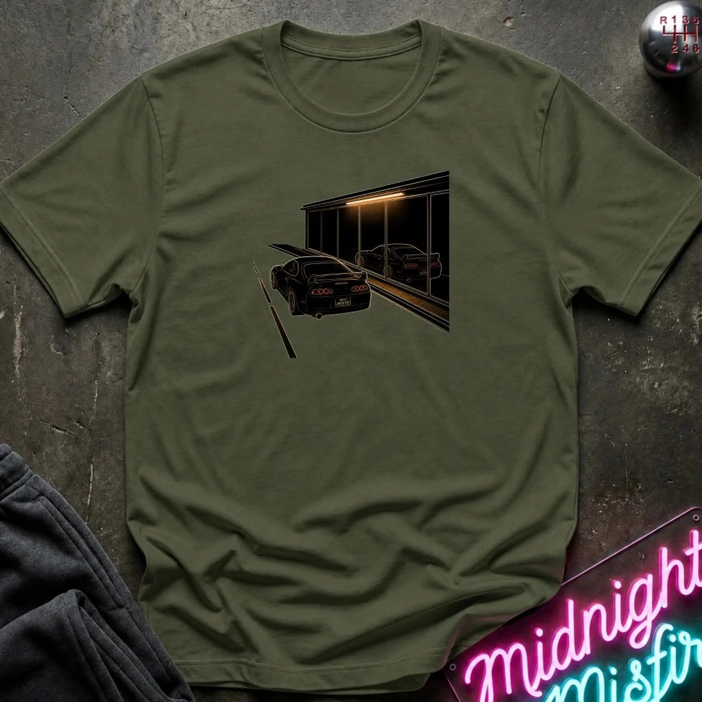 An olive green short-sleeve t-shirt laid flat on a dark textured surface, featuring a graphic print of a black sports car in front of a dark building with warm light. 