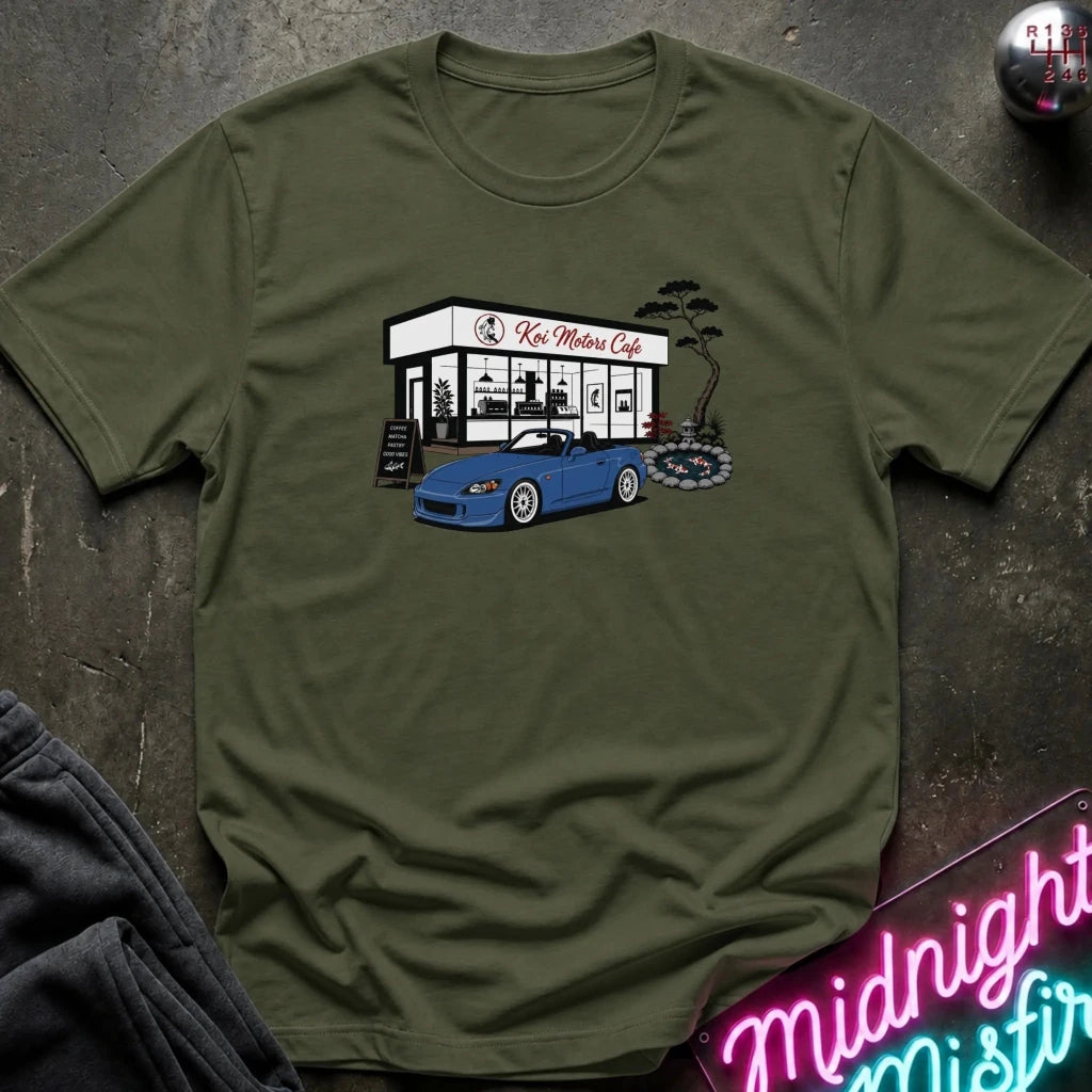 An olive green  short-sleeve t-shirt laid flat on a dark textured surface, featuring a graphic print of a blue sports car parked in front of 'Koi Motors Cafe' with a small tree illustration. 