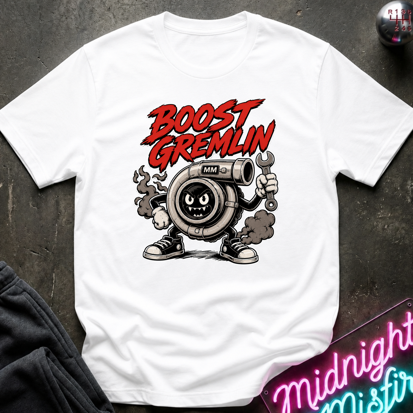 A white short-sleeve graphic T-shirt laid flat on a dark textured surface. The shirt features a cartoon turbocharger character with the words 'BOOST GREMLIN' in red above it.